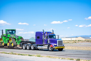 Oversize cargo hauler big rig purple classic semi truck tractor transporting oversize load on step deck semi trailer driving on the straight highway road