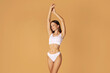 Young caucasian woman posing in white underwear with raised hands over peach studio background, lady showing her perfect ideal body shape, copy space