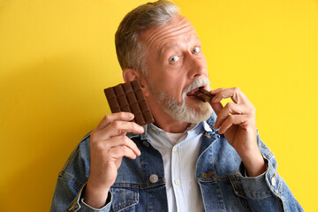 Mature man eating tasty chocolate on yellow background, closeup