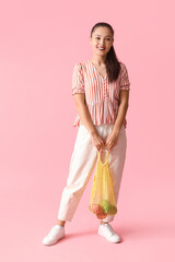 Poster - Young Asian woman holding strip bag with fresh apples on pink background