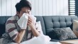 Sick young asian man sitting on sofa and sneeze with tissue paper at home. man blowing nose, coughing or sneezing in tissue at home, suffering from flu. Cold and fever concept.