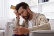 © New Africa - Upset man with credit card using smartphone in armchair at home. Be careful - fraud
