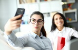 Fototapeta Nowy Jork - Portrait of beautiful businesswoman taking photo with colleague. Wonderful businesslady with black long hair wearing stylish glasses and posing for selfie. Business concept