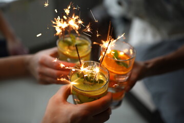 Wall Mural - Young group friends hold in hand fruit cocktail with sparkler decoration. Christmas invite party concept. Heath care vegan summer drinks