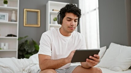 Poster - Young latin man using touchpad sitting on the bed wearing headphones at bedroom