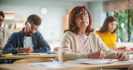 Leinwandbilder - Portrait of Happy Senior Woman Taking a Course in an International Adult Education Center. Old Female Wearing Glasses, Sitting Behind a Desk, Using Textbook and Writing Down Notes in Notebook