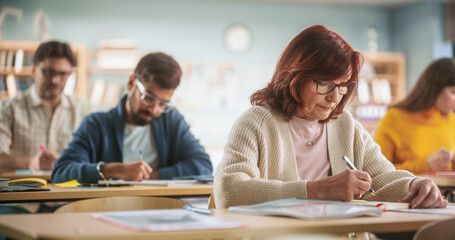 Fototapete - Portrait of a Happy Senior Woman Taking a Course in an International Adult Education Center. Old Female Wearing Glasses, Sitting Behind a Desk, Using Textbook and Writing Down Notes in Notebook