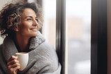 Fototapeta  - Portrait of happy middle aged woman in cozy sweater holding a cup of hot drink and looking trough the window, enjoying the winter morning at home, side view