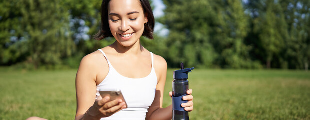 Wall Mural - Smiling fitness girl drinks water, checks her app on smartphone and looks happy, stays hydrated on fresh air, sunny day in park