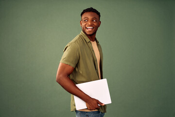 Wall Mural - Student using modern gadgets. Smiling african american man standing in half turn with wireless laptop and looking aside over green background. Happy guy in casual khaki shirt using portable computer.