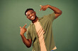 Perfect smile. Attractive african american guy wearing unbuttoned khaki shirt pointing with index fingers on his white teeth over green studio background. Concept of dental health.
