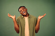 Oops, don't know. Pleasant african american guy shrugging shoulder and keeping palms wide open while standing over green background. Young man smiling with open mouth and expressing sincere emotions.