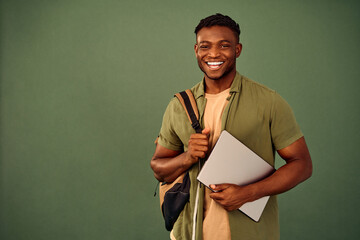 Distance learning. Glad african american guy with backpack and wireless laptop posing over green background. Enthusiastic smiling student engaging in online education with help of modern gadget.