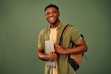 Wall Mural - Technology and e-learning. Portrait of enthusiastic afro student looking at camera with smile while holding laptop. Happy young guy standing over green background and carrying backpack on shoulder.