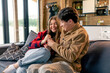© Guys Who Shoot - young couple in love fooling around, laughing and fighting over TV remote control while sitting on sofa