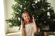 A girl with brown long hair in a white T-shirt and red plaid pants under a Christmas tree in a room for the New Year at home opens gifts with headphones, dresses and listens to music.