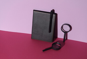 Poster - Notebook with black leather cover and pen, magnifying glass on pink background. Creative layout. Business concept