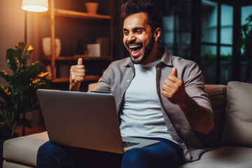 Handsome young latino man smiling and rejoicing after success. Happy man celebrating business success on sofa in living room with computer.