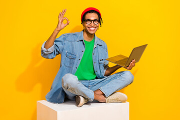 Full length photo of cute young male sitting white platform show okey laptop wear trendy jeans garment isolated on yellow color background