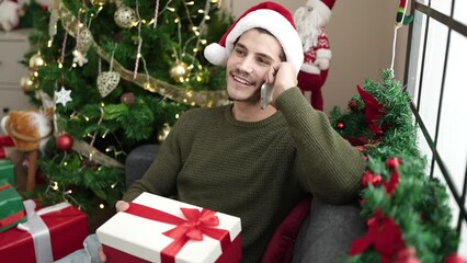 Wall Mural - Young hispanic man talking on smartphone sitting on sofa holding christmas gift at home