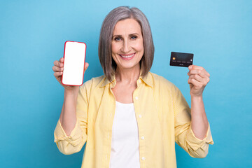 Poster - Photo of charming cheerful grandmother hold showing empty space smart phone display debit card isolated on blue color background