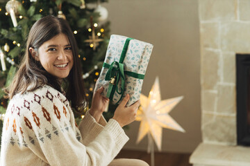 Wall Mural - Woman in cozy sweater holding stylish christmas gift on background of decorated christmas tree with lights in living room. Atmospheric winter holidays time. Preparing Christmas presents