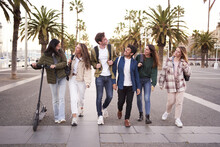 Diverse multicultural group young millennial friends walking along urban street palm trees. University people happy strolling outside on way to campus. Concept of cheerful students together. 