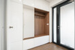 Modern entrance room of a family house with built-in wardrobes and a wardrobe wall made of white material and wood. The entrance door is glazed with frosted glass. The floor is wooden.