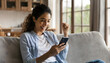 Happy excited young latin woman relaxing on couch using phone winning money in online app 