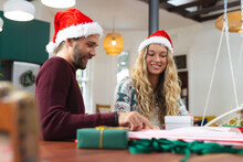 Happy Caucasian Couple In Christmas Hats Wrapping Presents At Kitchen Table, Copy Space