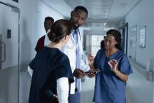 Diverse Male And Female Doctors Discussing Work, Using Tablet In Corridor At Hospital