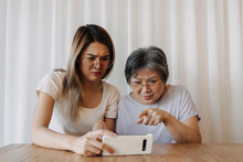 Asian Thai Old Mother And Daughter Using Phone Together With Doubt Face, One Holding And One Pointing Finger At Tablet, Curios And Surprised With Something On Screen.