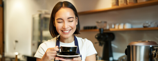 Close up of smiling asian girl barista, worker in cafe, drinking coffee on her break, holding cup with pleased face and closed eyes, enjoying her drink