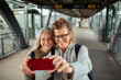 © Marko Geber - Two senior female friends taking selfies on a smartphone at the train station