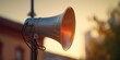 A silver megaphone is pictured on a pole with a building in the background. This image can be used to represent communication, announcements, or public speaking events.
