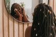 Happy plus size African woman cleaning face with cotton pad and looking at the mirror in bathroom