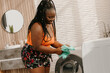 © gstockstudio - Beautiful plus size African woman loading clothes into washing machine while standing in domestic bathroom