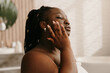 © gstockstudio - Confident curvy African woman covered in towel applying hydrating face cream in bathroom