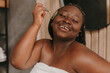 © gstockstudio - Beautiful plus size African woman covered in towel massaging face with roller at the domestic bathroom