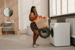 © gstockstudio - Happy plus size African woman carrying basket with clothes while standing near the washing machine in bathroom