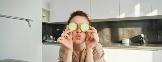 Wall Mural - Funny, happy girl eating healthy, holding zucchini, chopping vegetables for healthy meal in the kitchen, posing in bathrobe