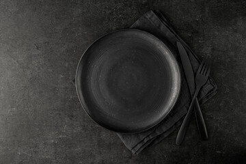 Empty grunge dark grey craftmade ceramic plate with cutlery and linen serviette on black stone board background. Retro style table