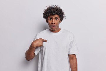 Wall Mural - Waist up shot of confused curly haired Hindu man points at himself and has puzzled expression asks who me being stunned to be mentioned or chosen dressed in casual basic t shirt isolated on white wall