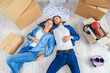 © gorynvd - Happy young caucasian couple lying down on the wooden floor while having a break while moving to new house. Top view.