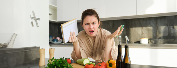 Wall Mural - Portrait of woman with angry face, standing near vegetables and looking frustrated, holding notebook, annoyed while cooking meal