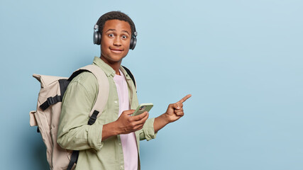 Horizontal shot of surprised dark skinned man holds mobile phone connected to headphones points finger at mock up space listens music via headphones isolated over blue backgound. Look at this