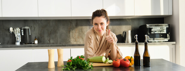 Wall Mural - Healthy lifestyle. Young woman in bathrobe preparing food, chopping vegetables, cooking dinner on kitchen counter, standing over white background