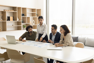 Wall Mural - Serious young project manager woman explaining sales marketing colleagues, investors, standing at meeting table, pointing at document with statistic data, speaking to partners