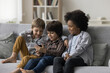 © fizkes - Three smiling cute boys sit on sofa in living room use mobile phone play online games on smartphone, watch funny video, friends spend free time together at home chatting with mates. Modern technology