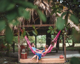 Fototapeta  - casal jovem deitado em rede cor de rosa em frente a uma maloca com teto de palha rodeado de árvores verdes, em Soure, ilha do Marajó 